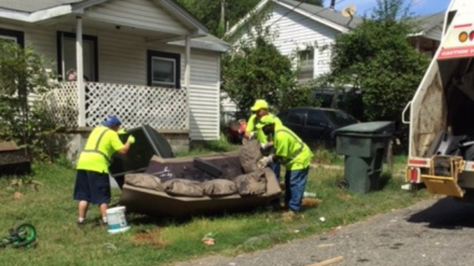 Crews in yellow vests lifting an old couch into a garbage truck